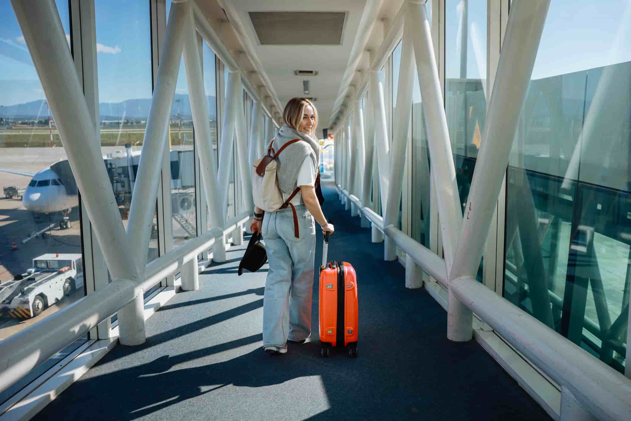 Woman with backpack and orange carry-on suitcase walking through a sunlit airport boarding bridge, with planes and apron visible through large windows