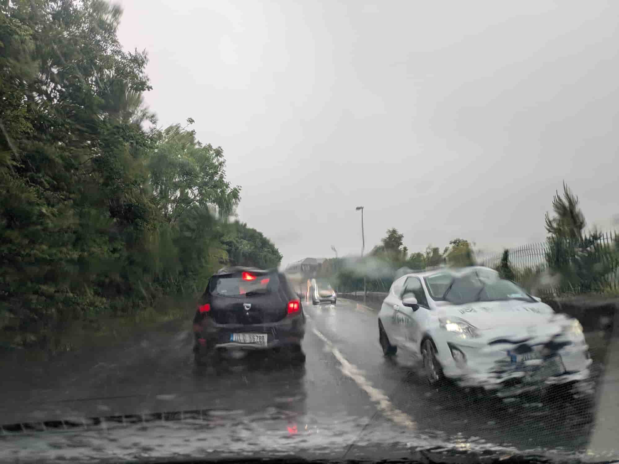 Two cars driving on a wet Irish road in heavy rain with reduced visibility and spray from the road surface.